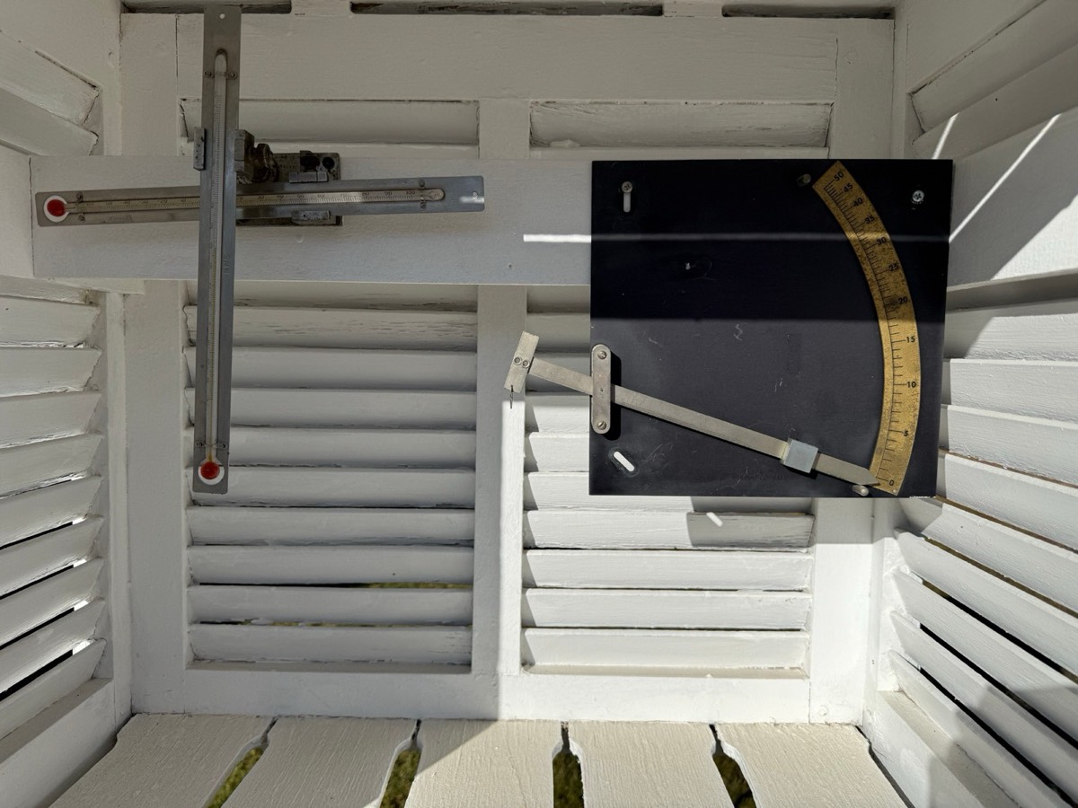 Inside the shelter: crossed maximum and minimum thermometers on the left, and a black fuel-moisture scale with a brass dial on the right